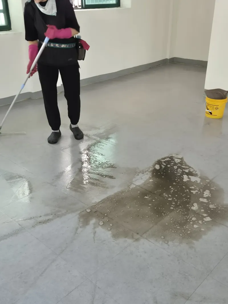 회색 근무복을 입은 작업자가 문턱 매트 위에 쌓인 포장재 부스러기를 손 빗자루로 쓸어 담는 모습, 뒤편 철제 방화문 너머로 팔레트가 쌓인 창고 통로가 보인다