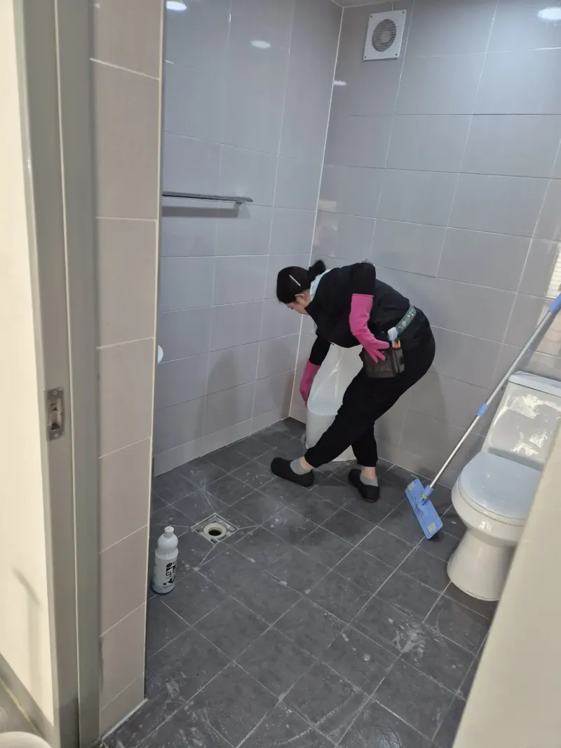 같은 화장실 칸에서 작업자가 허리를 숙여 소변기 안쪽을 정리하는 모습, 옆으로 양변기와 파란 손잡이 대걸레, 바닥의 세정제 통이 함께 보이는 장면